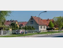 Straßenszene in einem Wohngebiet mit Einfamilienhäusern, die rote und orange Ziegeldächer haben. Im Vordergrund befinden sich ein Zaun, ein Stromkasten mit Dekoration, sowie eine kleine Gruppe von Kindern neben Fahrrädern; im Hintergrund stehen Bäume und Sträucher.