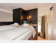 Modernes Schlafzimmer mit großem Bett und weißer Bettwäsche, integriert in eine dunkle, holzverkleidete Wandstruktur mit Ablage, Büchern und zwei Leuchten. Rechts befindet sich ein Fenster mit schweren Vorhängen sowie eine schmale Sitznische und ein Heizkörper an der Wand.