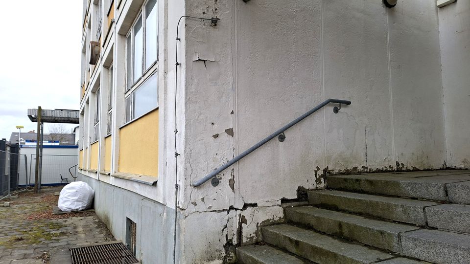 Außenansicht eines älteren Mehrgeschossgebäudes mit großen Fenstern und abblätterndem Putz an der Fassade. Rechts führt eine steinerne Treppe mit Metallhandlauf zu einem erhöhten Eingang, daneben ein gepflasterter Weg entlang der Gebäudeseite.