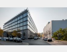 Moderne Bürogebäude mit großen Glasfassaden und klaren Linien an einer Straßenecke, umgeben von geparkten Autos, Herbstbäumen und Gehwegen. Rechts daneben steht ein schlichtes graues Gebäude mit glatter Fassade.