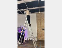 Interior of an open room with a high ceiling, exposed brick wall, and dark acoustic ceiling panels on a metal frame. A tall aluminum step ladder stands on a wood floor marked with tape, with cables and small hanging decorations near the ceiling.