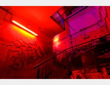 Eine urbane Treppe in einem Innenraum, beleuchtet von roten und violetten Neonlichtern. Die Wände sind mit Graffiti und sichtbaren Rohren sowie Kabeln bedeckt, während Metallgeländer die Treppenabsätze einfassen.