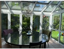 Wintergarten mit gläsernem Dach und großen Fenstern, die den Blick auf einen sonnigen Garten mit Bäumen und Blumen bieten. Im Raum steht ein runder Esstisch mit unterschiedlichen Stühlen, darüber hängt ein Kronleuchter.