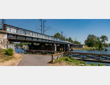 Bahnbrücke über einen Fluss mit Graffiti an den Brückenwänden, umgeben von Bäumen und einem Fußweg mit Geländer im Vordergrund. Im Hintergrund sind Strommasten und ein kleiner Bootsanleger zu sehen.