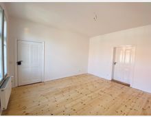 Leerer Raum mit hellen Holzdielen, weißen Wänden und zwei weißen Türen. Links im Bild befindet sich ein Fenster mit Heizkörper darunter.