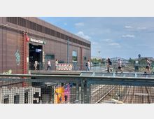 Bahnhof Warschauer Straße in Berlin, mit moderner, dunkelbrauner Fassadenverkleidung und Eingangsbereich zur S-Bahn. Im Vordergrund führt eine Fußgängerbrücke über mehrere Bahngleise, auf der sich Menschen bewegen; im Hintergrund sind Stadtansichten mit einer Brücke und einem Kran zu sehen.