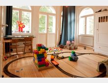 Ein helles Kinderzimmer mit Holzboden, großen Fenstern und blauen Vorhängen. Der Raum ist mit Spielzeug ausgefüllt, darunter bunte Bauklötze, eine Spielzeugeisenbahn, ein Modell einer Ritterburg und ein Kindertisch mit Stühlen.