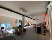 Großer, offener Raum mit industriellem Charme, weißen Säulen und Balken, hohen Fenstern mit Rundbögen, und minimalistischer Einrichtung. Im Raum befinden sich Sitzbereiche mit Teppichen, ein Gitternetzbehälter, Kamerastative sowie Stehtische, beleuchtet durch natürliches Licht und vereinzelte warme Akzentbeleuchtung.