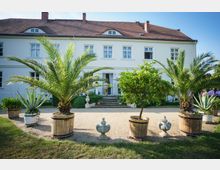 Ein zweistöckiges weißes Gebäude mit Satteldach, davor ein Garten mit Palmen, Orangenbäumen und Agaven in hölzernen Pflanzkübeln sowie einer dekorativen steinernen Vase auf einem Kiesweg. Im Hintergrund führen Treppenstufen zum Hauseingang, und Menschen sind zu sehen.