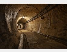 Ein unterirdischer Backsteintunnel mit gewölbter Decke, beleuchtet durch mehrere Leuchtstofflampen. Im Tunnel verläuft ein gepflasterter Weg mit Schienen, und an den Wänden sind Rohre und Kabel befestigt.