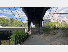 Ansicht eines Fußgängerwegs unter einer erhöhten Brücke mit Stahlkonstruktion, flankiert von Geländern und Grünflächen. Im Hintergrund sind mehrspurige Straßen, Ampeln, Wohngebäude und Bäume zu sehen.