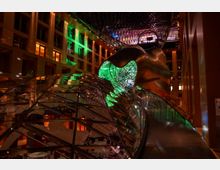 Beleuchtetes modernes Atrium mit einer geschwungenen, goldfarbenen Metallstruktur und gläsernen Fassadenelementen, durchsetzt von farbigen Lichteffekten in Grün und Orange. Eine geschwungene Rampe und eine filigrane Glasdecke prägen das futuristische Design des Raums.