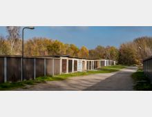 Reihen von Garagen mit flachen Dächern und unterschiedlich farbigen Toren, entlang eines unbefestigten Weges. Im Hintergrund befinden sich Bäume mit herbstlichem Laub und ein klarer Himmel.