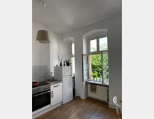 Helle Küche in einer Altbauwohnung mit hohen Decken, weißen Wänden und einem Holzboden. Die Ausstattung umfasst eine moderne Einbauküche, einen weißen Kühlschrank und ein geöffnetes Altbaufenster mit Blick auf grüne Baumkronen; darunter befindet sich eine Heizung.