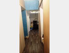 Ein schmaler Flur mit dunklem Laminatboden, flankiert von Schränken auf beiden Seiten. Am Ende des Flurs befindet sich ein kleiner Arbeitsplatz mit einem Schreibtisch, einem Computer und einer Lampe; darüber ein helles Oberlicht.