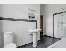 Modernes Badezimmer mit weißen Wandfliesen und dunklem Fliesenboden, ausgestattet mit Standwaschbecken, Toilette und rundem Spiegel unter einer Wandleuchte. Rechts ist eine Badewanne mit Glasduschwand sowie ein Fenster mit Sicht ins Grüne zu sehen.