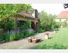 Gartenansicht eines ländlichen Anwesens mit gepflastertem Weg, umgeben von Blumenbeeten mit lila Blüten, Büschen und einem Gebäude mit gelber Fassade und roten Dachziegeln. Im Vordergrund stehen Körbe mit Holz und eine Feuerstelle.