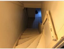 Holztreppe in einem engen Treppenhaus mit abblätterndem weißen Lack, die in einen gefliesten Untergeschossbereich führt. Am unteren Ende des Treppenabsatzes sind Schuhe auf dem Boden sichtbar.