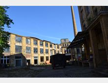 Ein verlassener Industriehof mit alten, gelblich-braunen Backsteingebäuden, von denen viele zerbrochene Fenster haben; im Hintergrund ragt ein hoher Schornstein empor. Im Vordergrund steht ein schwarzes Fahrzeug, und der Boden ist mit Kopfsteinpflaster und Beton bedeckt.