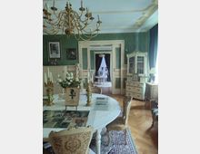 Elegantes Wohnzimmer im historischen Stil mit grünen Wänden und weiß-goldener Zierde, einem runden Tisch mit Spitzendecke und Dekorationen. Im Hintergrund sind ein Durchgang mit Doppeltüren sowie ein angrenzender Raum mit Vorhängen und einer Kommode sichtbar.