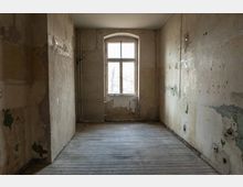 Ein leerer, heruntergekommener Raum mit abgenutzten Holzdielen, beschädigten Wänden mit abgeblätterter Farbe und einem großen, bogenförmigen Fenster, das diffuses Tageslicht hereinlässt.