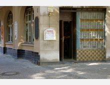 Straßenszene mit der Fassade eines Gebäudes, das einen Eingang zu einem Lokal zeigt. Der Eingang ist umgeben von Glasbausteinen mit gelben Akzenten, einer offenen Tür und einem beleuchteten Schild mit der Aufschrift „Schultheiss“. Links befinden sich Fenster mit Rundbögen und dunklen Fliesen an der unteren Wand.