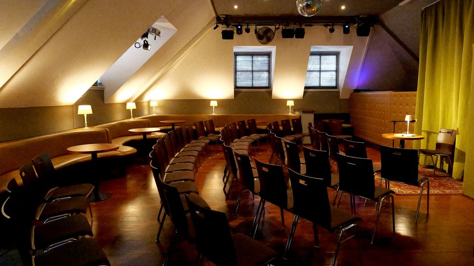 Dachgeschoss-Veranstaltungsraum mit schrägen Decken, Holzfußboden und bogenförmig angeordneten Stuhlreihen vor einer kleinen Bühnenfläche mit gelbem Vorhang. Entlang der Wand stehen gepolsterte Sitzbänke mit kleinen runden Tischen und Tischlampen; an der Decke hängen Scheinwerfer und eine Diskokugel, hinten sind zwei Fenster.