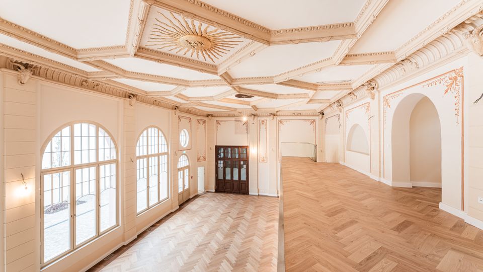 Heller, leerer Innenraum mit großen bogenförmigen Sprossenfenstern, Fischgrätparkett und einer erhöhten Galerieebene. Die Decke ist reich mit Stuck und einem zentralen Sonnenmotiv verziert, dazu helle Wandpaneele und Rundbogenöffnungen.