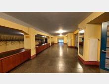 Ein großzügiger Garderobenbereich mit gelb gestrichenen Wänden und roten Sockeln, langen hölzernen Ablagen und einer Reihe von Kleiderhaken an Metallgestellen. Der Raum verfügt über rechteckige Säulen, eine Spiegelwand, Heizkörper und eine blaue Tür auf der rechten Seite.