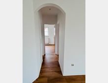 Schmaler, heller Wohnungsflur mit weiß verputzten Wänden, Holzboden und einem Rundbogen-Durchgang. Am Ende führt eine offene Tür in einen Raum mit Fenster und darunterliegendem Heizkörper.