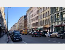 Straßenszene in einer Stadt mit mehrstöckigen Altbauten, Geschäften im Erdgeschoss und parkenden Autos am Straßenrand. Eine belebte Fahrbahn mit Fahrzeugen sowie ein heller, sonniger Tag zeichnen die Umgebung aus.