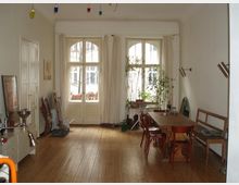 Wohnzimmer mit Holzboden, einem langen Holztisch und Stühlen auf der rechten Seite, großen Fenstern mit weißen Vorhängen im Hintergrund und einzelnen Pflanzen als Dekoration. Links stehen weitere Möbel und Gegenstände, während die Raumdecke mit bunten Hängelichtern geschmückt ist.