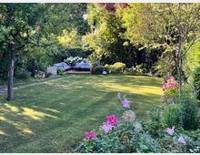 Ein gepflegter Garten mit einer Rasenfläche im Vordergrund, umgeben von bunten Blumenbeeten und Bäumen. Im Hintergrund befindet sich eine kleine Terrasse mit einer Sitzecke aus Gartenmöbeln, dekoriert mit Kissen und umgeben von üppigem Grün.
