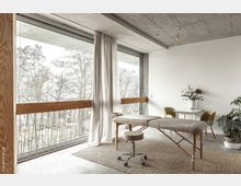 Heller Raum mit großen bodentiefen Fenstern, die den Blick auf Bäume freigeben. Im Vordergrund stehen eine Massagebank und ein Hocker, im Hintergrund ein kleiner Tisch mit zwei Stühlen und einer Blumenvase.