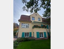 Gelbes Wohnhaus mit rotem Ziegeldach und grünen Fensterläden, umgeben von einem gepflegten Garten. Ein Balkon mit Blumenkästen ragt über die untere Etage, und ein Baum mit sichtbarer Baumkrone rahmt das Bild teilweise ein.