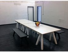 Ein schlichter, leerer Raum mit dunklem Boden und weißen Wänden. In der Mitte stehen zwei weiße Tische auf Holzböcken, flankiert von schwarzen Bänken; darauf steht eine Schale mit Obst. Im Hintergrund sind zwei schlichte Türen sichtbar.