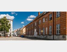 Straßenszene mit einem historischen roten Backsteingebäude, das große weiße Fenster und goldene Verzierungen besitzt, sowie einem Café unter einem Baum auf der linken Seite. Rechts führt eine schmale Straße entlang moderner und älterer Gebäude bei klarem Himmel mit vereinzelten Wolken.