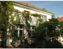 Zweistöckiges Wohnhaus mit weißer Fassade und roten Ziegeln auf dem Dach, teilweise von grünen Kletterpflanzen und Sträuchern umgeben. Das Gebäude hat große Fenster mit braunen Rahmen und steht in einer Gartenumgebung bei sonnigem Wetter.