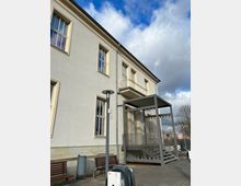 Außenansicht eines hell verputzten Gebäudes mit großen rechteckigen Fenstern und einem kleinen Balkon an der ersten Etage. Im Vordergrund befinden sich Sitzbänke, eine Laterne und eine Ladestation für Elektrofahrzeuge. Ein moderner Metalltreppenaufgang mit Überdachung ist an der Gebäudeseite angebracht.