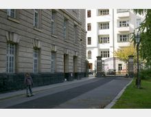 Straßenansicht mit einem Gebäude aus Steinfassade und vergitterten Fenstern auf der linken Seite, einem dekorativen Metalltor in der Mitte und einem hellen Wohngebäude mit Balkonen im Hintergrund; ein Kind fährt auf einem Skateboard entlang des Gehwegs.