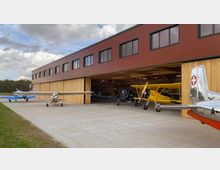 Moderne Flugzeughalle mit Holzverkleidung und geöffnetem Tor, in der mehrere kleine Flugzeuge, darunter eine gelbe Doppeldeckermaschine, geparkt sind. Im Vordergrund befinden sich weitere Kleinflugzeuge auf einer gepflasterten Fläche, und im Hintergrund ist ein bewölkter Himmel zu sehen.