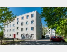 Ein Wohngebäude mit vier Etagen und schlichter grauer Fassade, umgeben von Bäumen und parkenden Autos. Im Vordergrund ist eine gepflasterte Straße mit schattenspendenden Bäumen zu sehen.