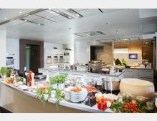 Moderne Kochschule mit großen Arbeitsflächen, voll ausgestatteten Küchenzeilen und integrierten Öfen. Auf der zentralen Theke befinden sich verschiedene Kochutensilien, Töpfe, frische Zutaten wie Kräuter, Gemüse und ein großer Parmesan-Laib.