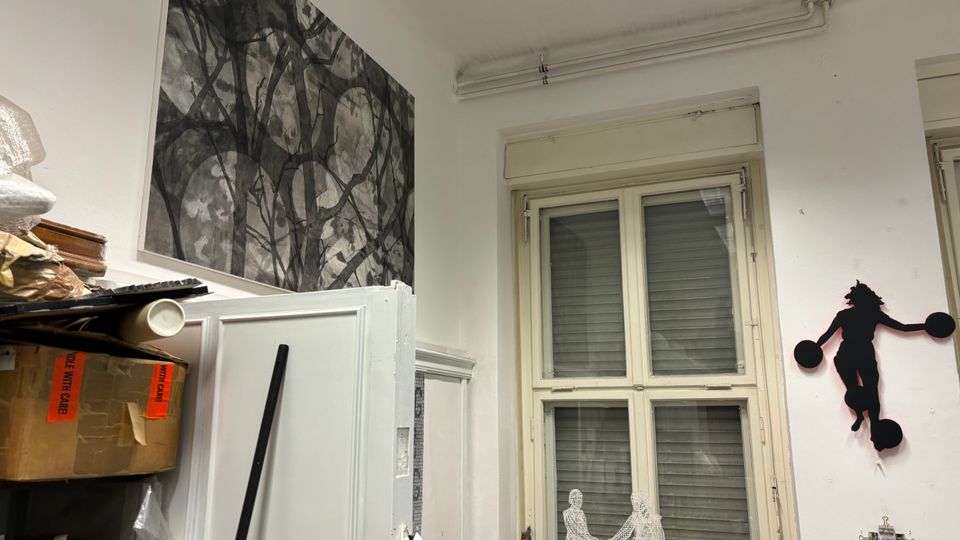 Innenraum eines Ateliers oder Lagerraums mit hohen weißen Wänden, großem Fenster mit geschlossenen Rollläden und sichtbaren Leitungen an der Decke. An den Wänden hängen Kunstwerke und Silhouetten, daneben stehen Schränke und gestapelte Kartons und Materialien.