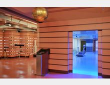 Ein Ausstellungsraum mit orangefarbenen Wänden und horizontalen Streifen, ausgestattet mit wissenschaftlichen Objekten wie Teleskopen und einer Raketen-Nachbildung. Im Hintergrund führt eine Tür mit blauem Licht in einen Bereich mit einer Raumkapsel-Nachbildung und Laufband.