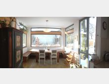 Wohnzimmer mit großem Fensterbereich, der den Blick auf einen verschneiten Garten bietet. Ein Holztisch mit weißen Stühlen und Sitzbänken steht in der Mitte, umgeben von hellen Wänden, Pflanzen und einem rustikalen Holzschrank links.