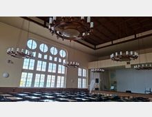 Großer Saal mit hohen, rundbogigen Sprossenfenstern und Holzbalkendecke, von mehreren ringförmigen Kronleuchtern beleuchtet. Im Vordergrund stehen viele Reihen von Tischen und Stühlen, hinten befindet sich eine Bühne mit Vorhang und Lautsprechertechnik.