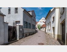 Schmale gepflasterte Gasse in einem historischen Wohnviertel mit hell verputzten Häusern, teils mit Fensterläden, und roten Ziegeldächern. Im Hintergrund sind weitere Gebäude und ein kleiner Baum sichtbar, bei klarem blauem Himmel.