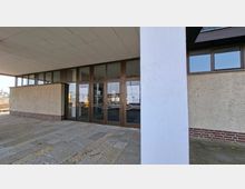 Eingangsbereich eines modernen Gebäudes mit überdachtem Vorplatz, breiten Steinplatten und einer großen weißen Stützsaule im Vordergrund. Dahinter befinden sich mehrere braune Glastüren mit seitlichen Fensterfronten und verputzten Außenwänden mit Sockel aus rotem Ziegelstein.
