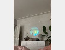 Ein schlicht eingerichtetes Zimmer mit einer weißen Kommode, auf der Pflanzen, eine Lampe und Dekorationsgegenstände stehen. Über der Kommode hängt ein runder Spiegel, der ein Fenster mit buntem Lichtreflexionen zeigt; daneben befinden sich große grüne Zimmerpflanzen.